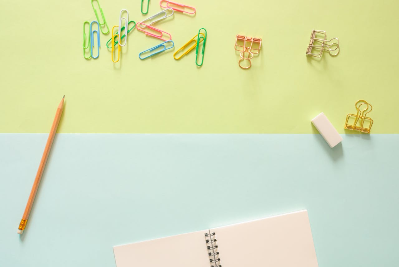 Top view of colorful stationery items, including a notebook, pencil, paperclips, and an eraser on pastel background.